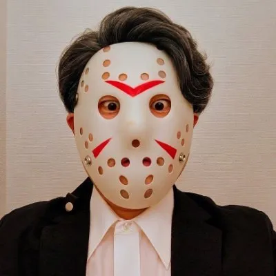 白い壁の前で、ジャケットに白いシャツを着て、目線の高さから撮った写真。顔はお面で隠している